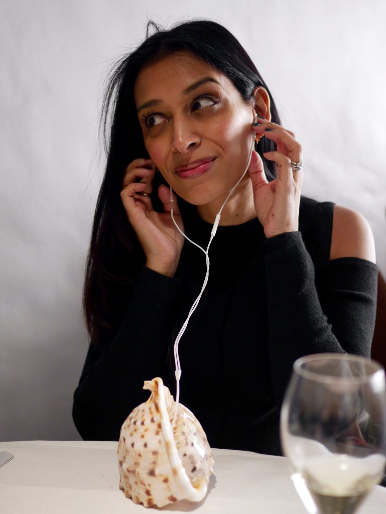 sima listening to earphones plugged into a conch shell playing a soundtrack of crashing waves and seagulls