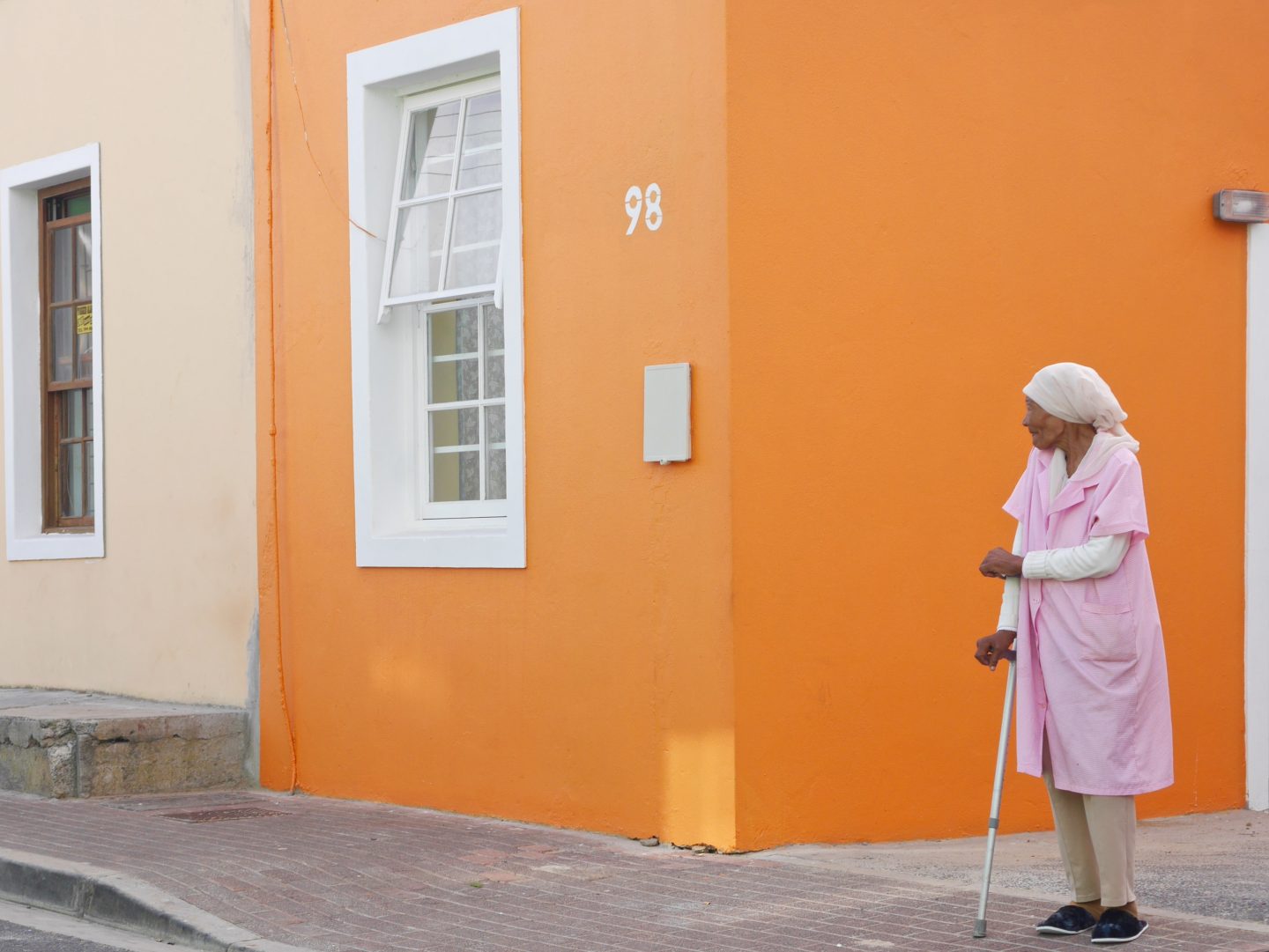 lady in pink with a walking stick at the side of a road against an orange house