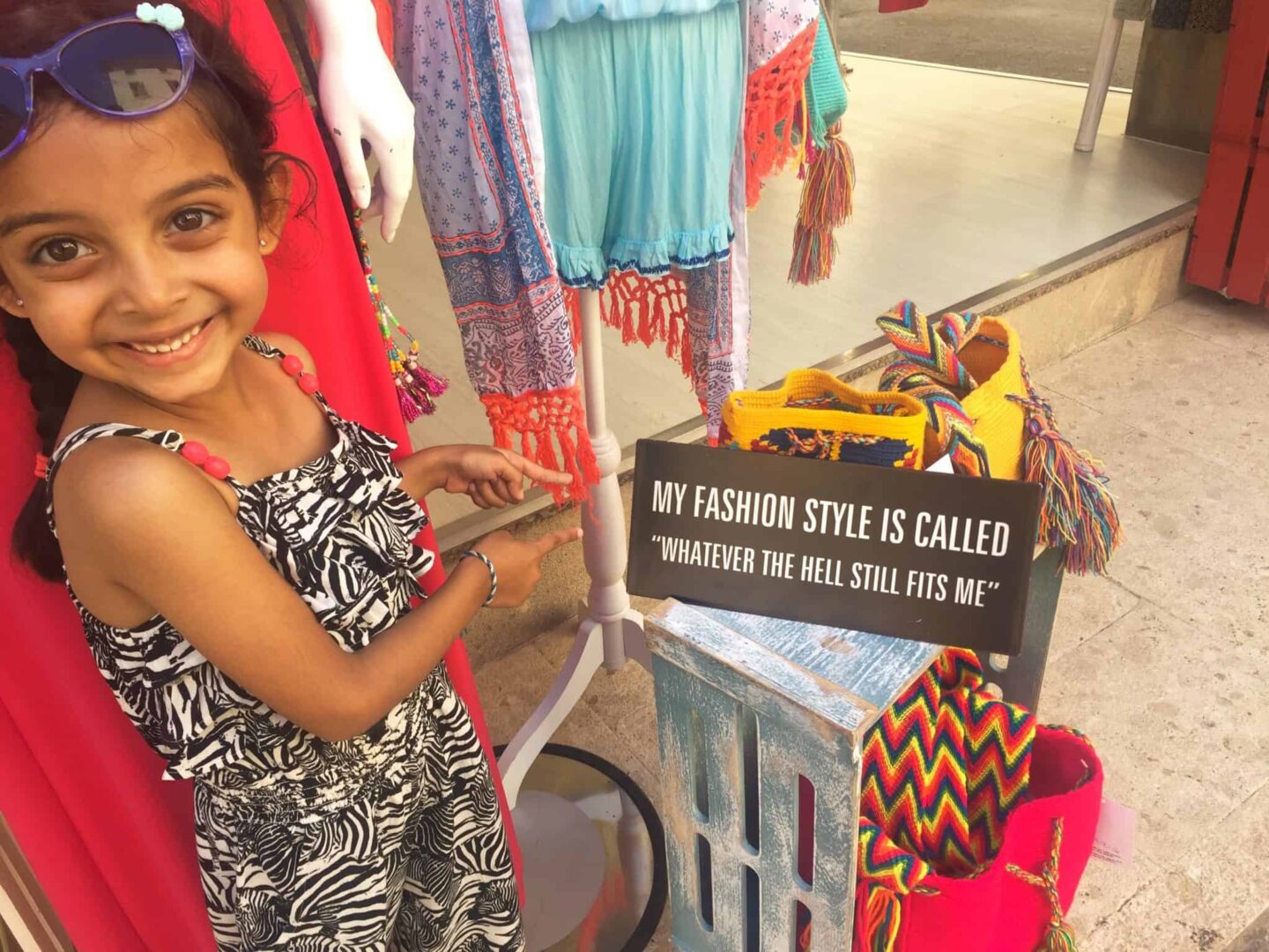 Daughter with blue glasses on head and zebra pattered jump suit pointing at a sign in a market 'my fashion style is called' 'whatever the hell still fits me'