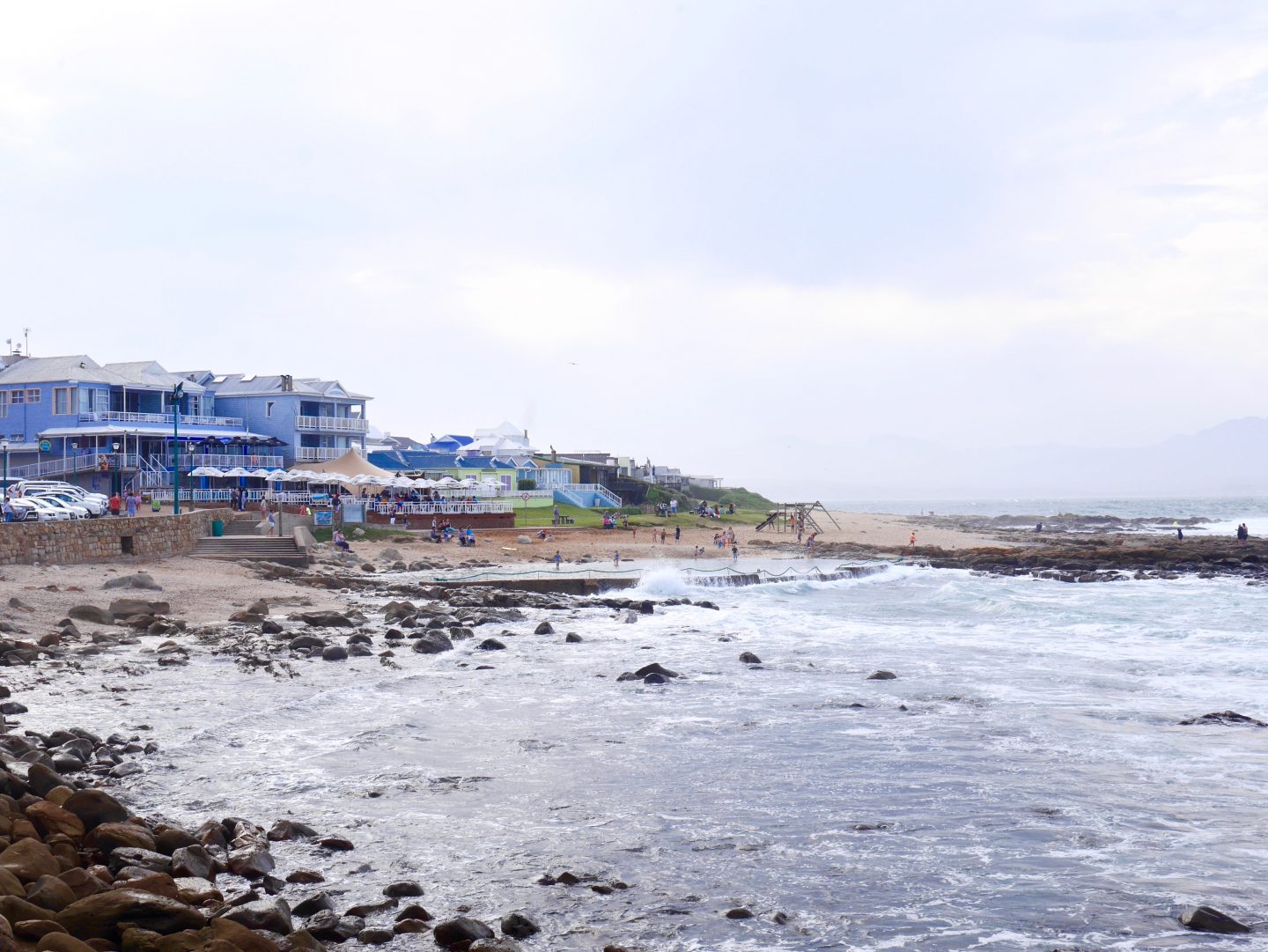 picture looking into the bay of Mossel Bay from the seat with waves crashing against large stones and breakers beneath a blue hotel with balconies and cars in carpark