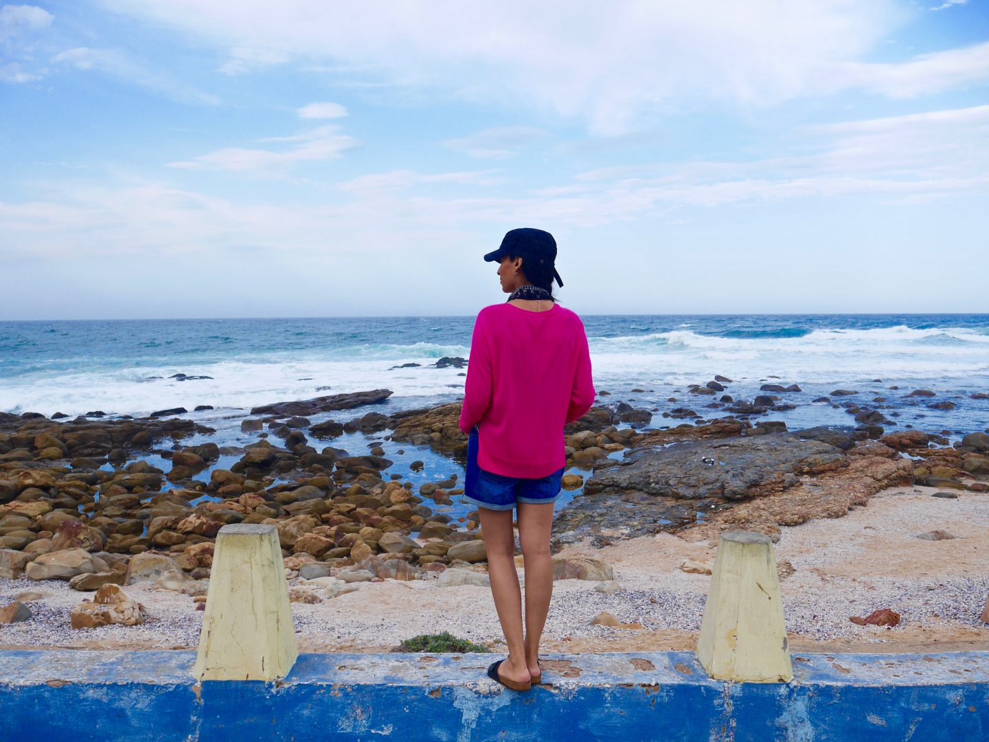 sima wearing pink top, denim shorts and black hat stood on a blue wall looking out to the sea surround by small amount of sand/gravel and mainly rocks between shore and sea