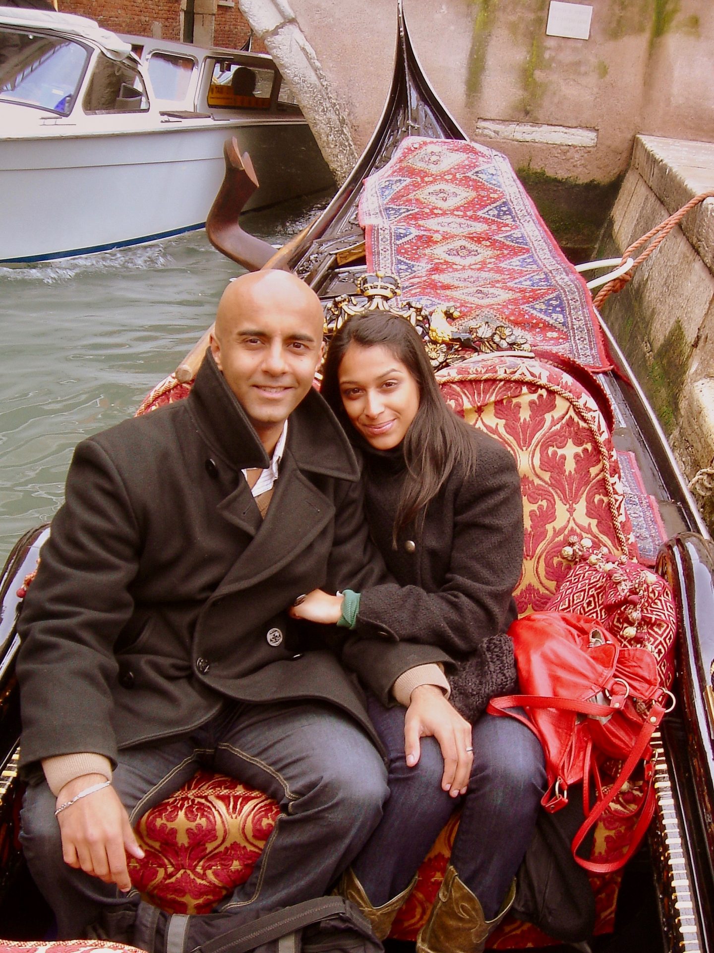 view of sima and husband wearing black coats in a gondola surrounded by red furnishings