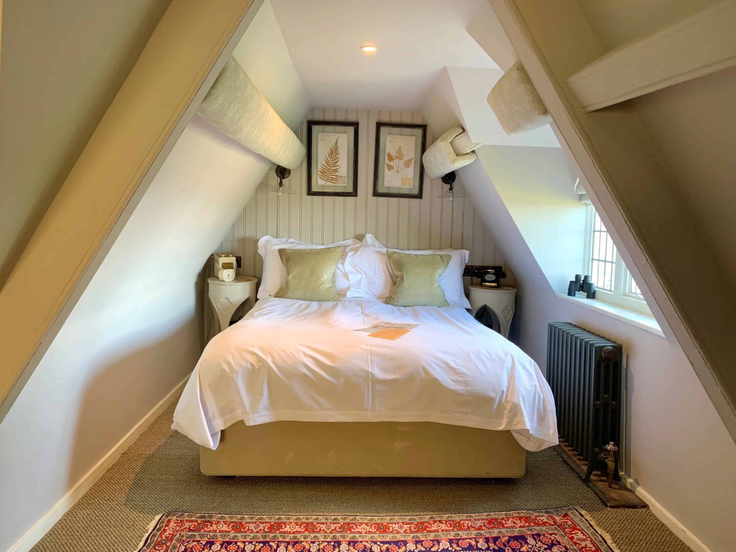 double bed in luxury Dorset hotel under sloping roof with window, white bed spread, green pillows, roof beams, striped paper and dark radiator