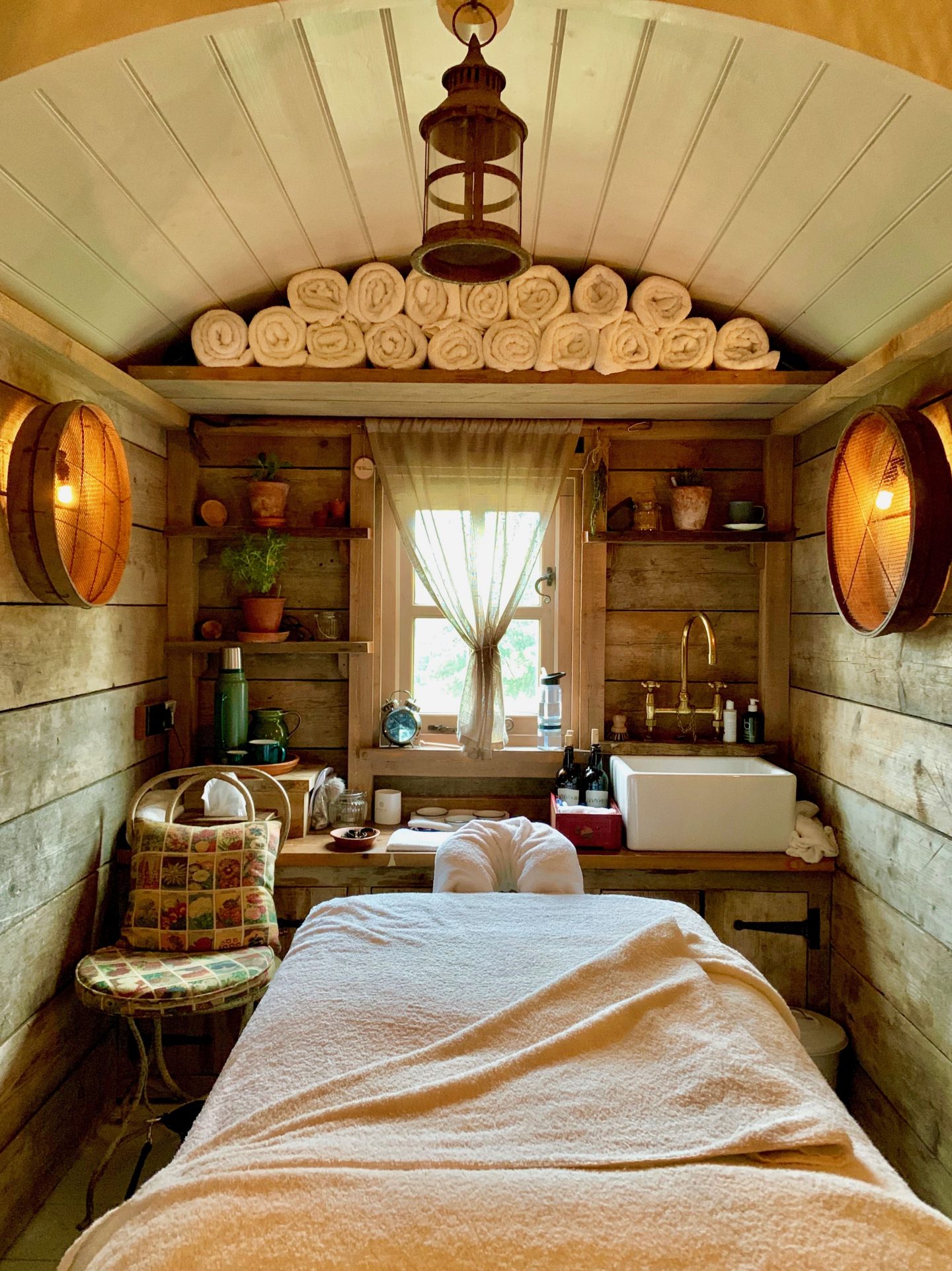 inside the wooden relaxation hut with warm lights, massage bed, small window, blind folded towels and a wicker chair