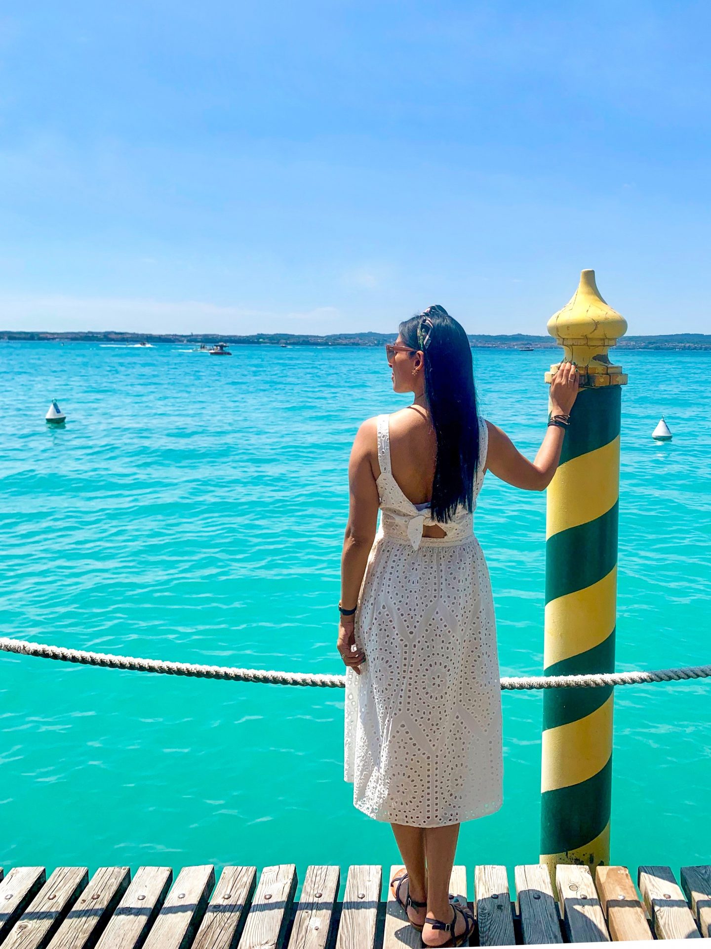 sima in white dress holding yellow and green pole behind rope on Lake Garda jetty overlooking the turquoise coloured sea