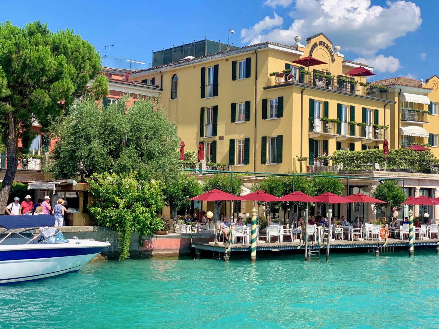 turquoise water with white and blue boat, cafe bars with red umbrellas and yellow buldings