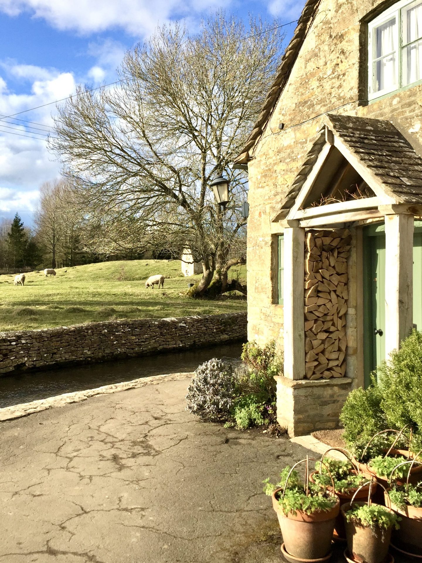 picture of a doorway with firewood outside the front door, nearby stream and sheep in The Cotswolds