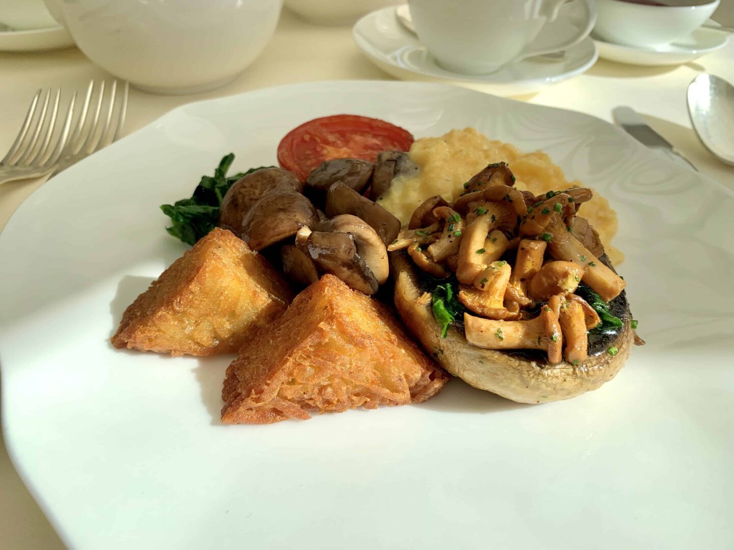 hot breakfast on a white plate with mushrooms, hash browns, scrambled egg and tomato.